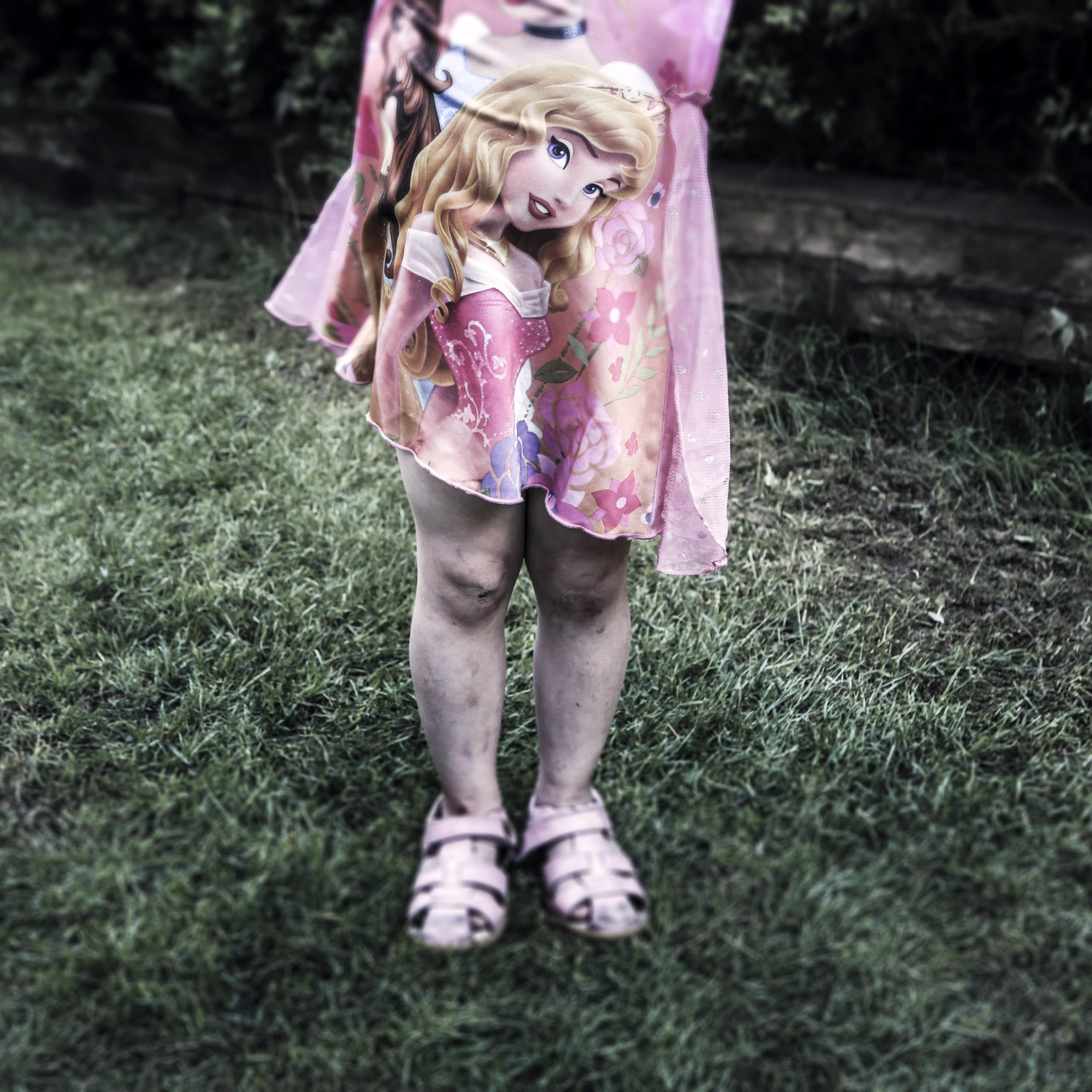 En liten jente i rosa kjole med står i gresset med søle på leggene. Vi ser henne fra livet og ned.