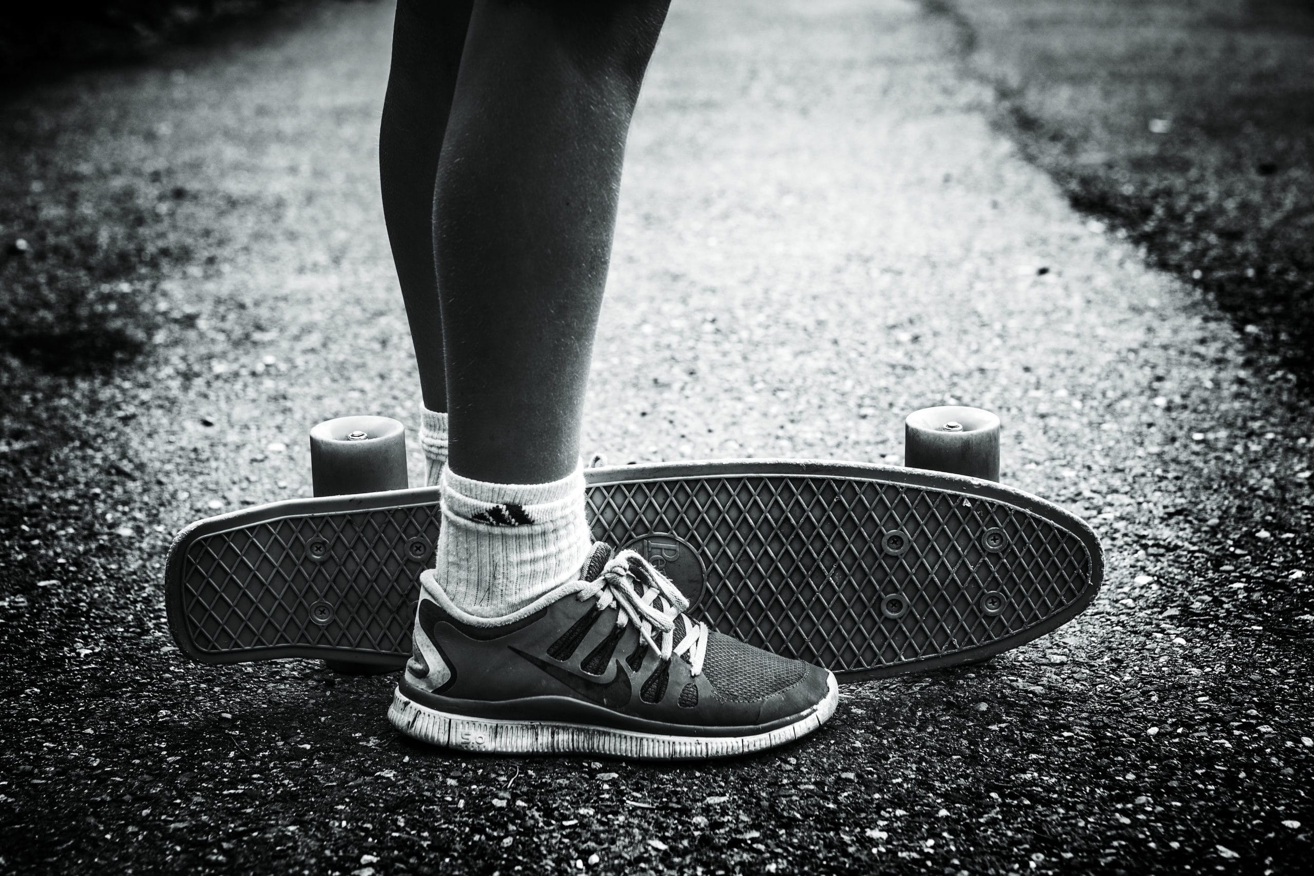 Svart/hvitt-bilde av to ben med joggesko med et skatebord i mellom.