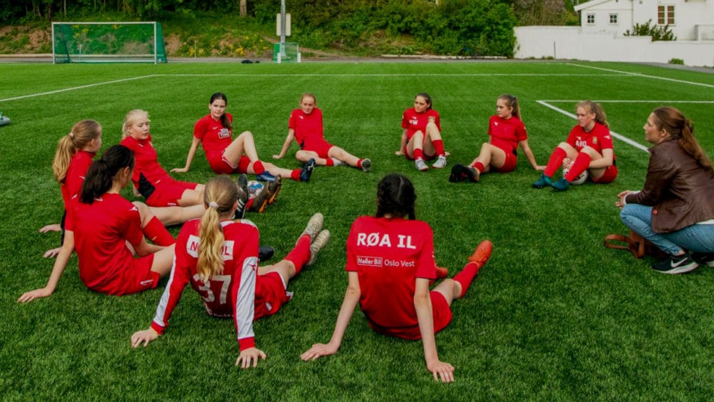 Fotballjenter sitter i ring på kunstgressbane sammen med en ansatt fra Redd Barna