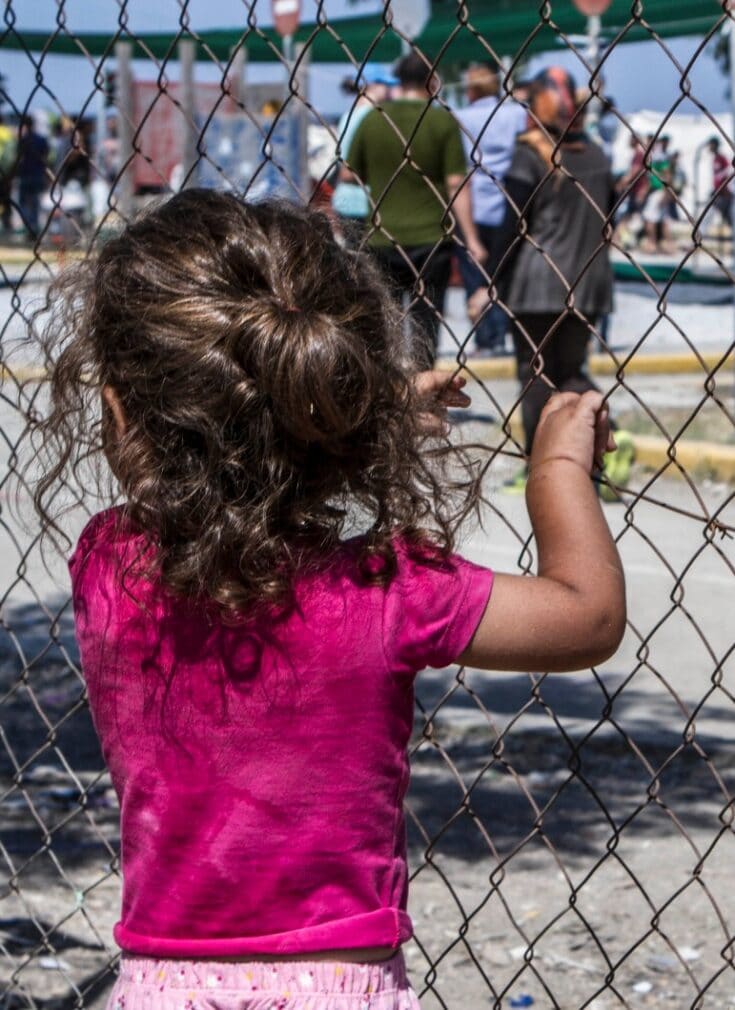 En liten jente står på innsiden av et gjerde i en flyktningeleirog ser ut.