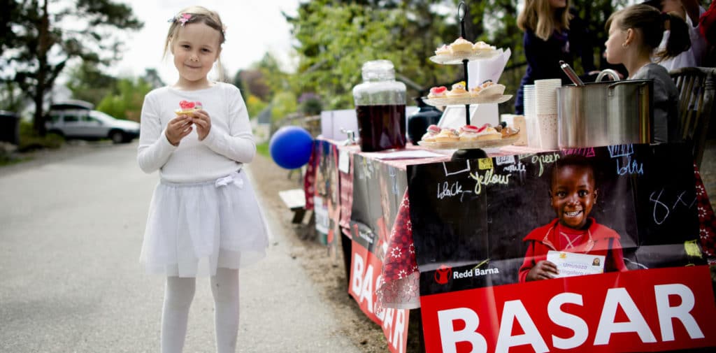 En jente står med en muffins i hånda ved siden av en basarbod med kaker og saft.