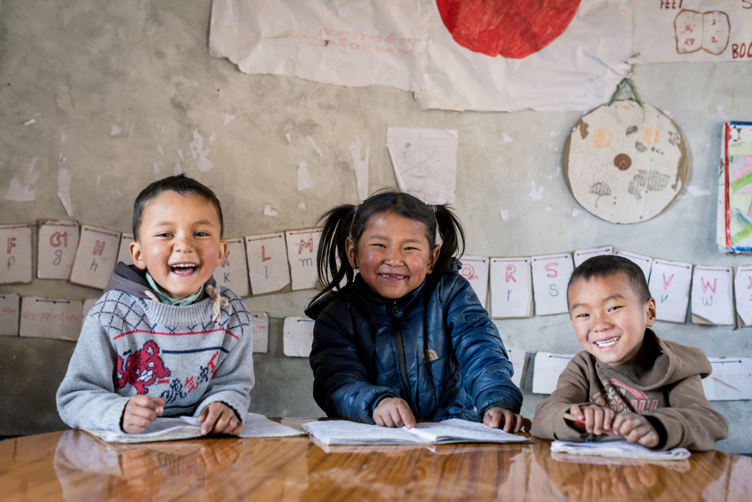 Tre skoleelever sitter med skolebøker foran seg og smiler bredt på Shree Buddha grunnskole i Dailekh, Nepal