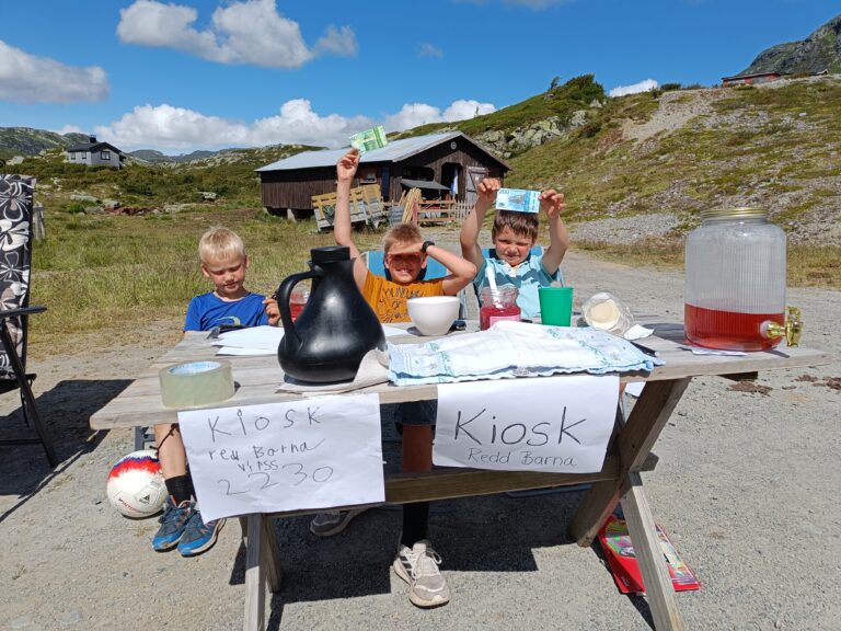 Tre gutter sitter ute ved sin hjemmelagede kiosk til inntekt for Redd Barna. De viser penger de har samlet inn.