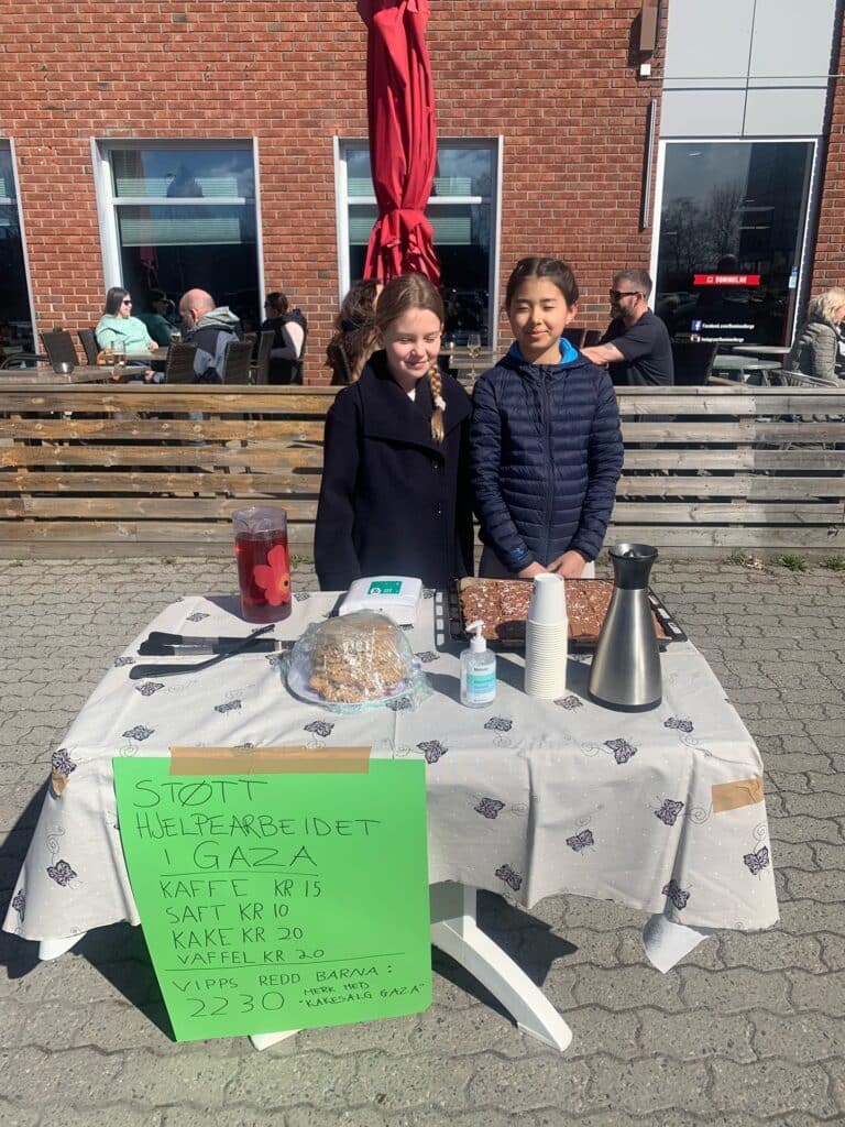 To jenter som smiler foran et bord, selger kake og kaffe