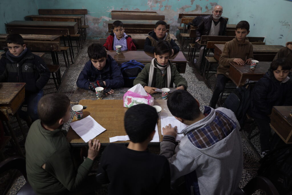 En gjeng barn sitter med jakker og skjerf på inne i et slitent klasserom i Syria