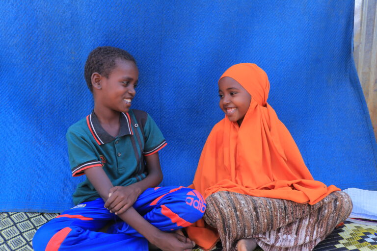 En gutt og ei jente sitter på gulvet og smiler og ser på hverandre