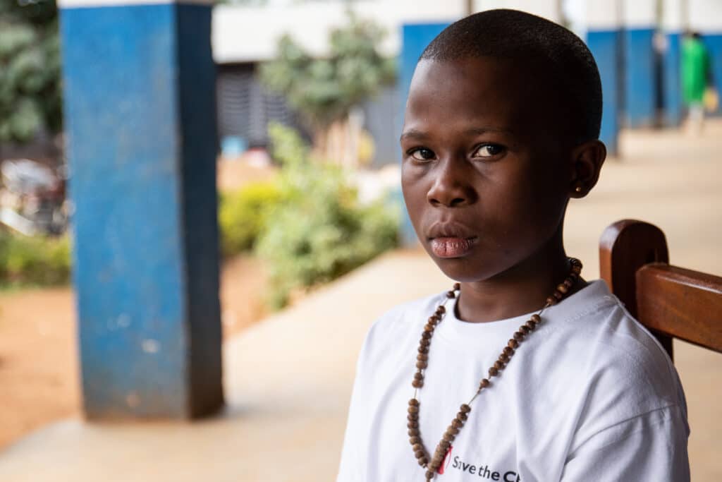 Ei jente i Redd Barna-t-skjorte sitter og kikker inn i kamera, utenfor en skole i Den demokratiske republikken Kongo