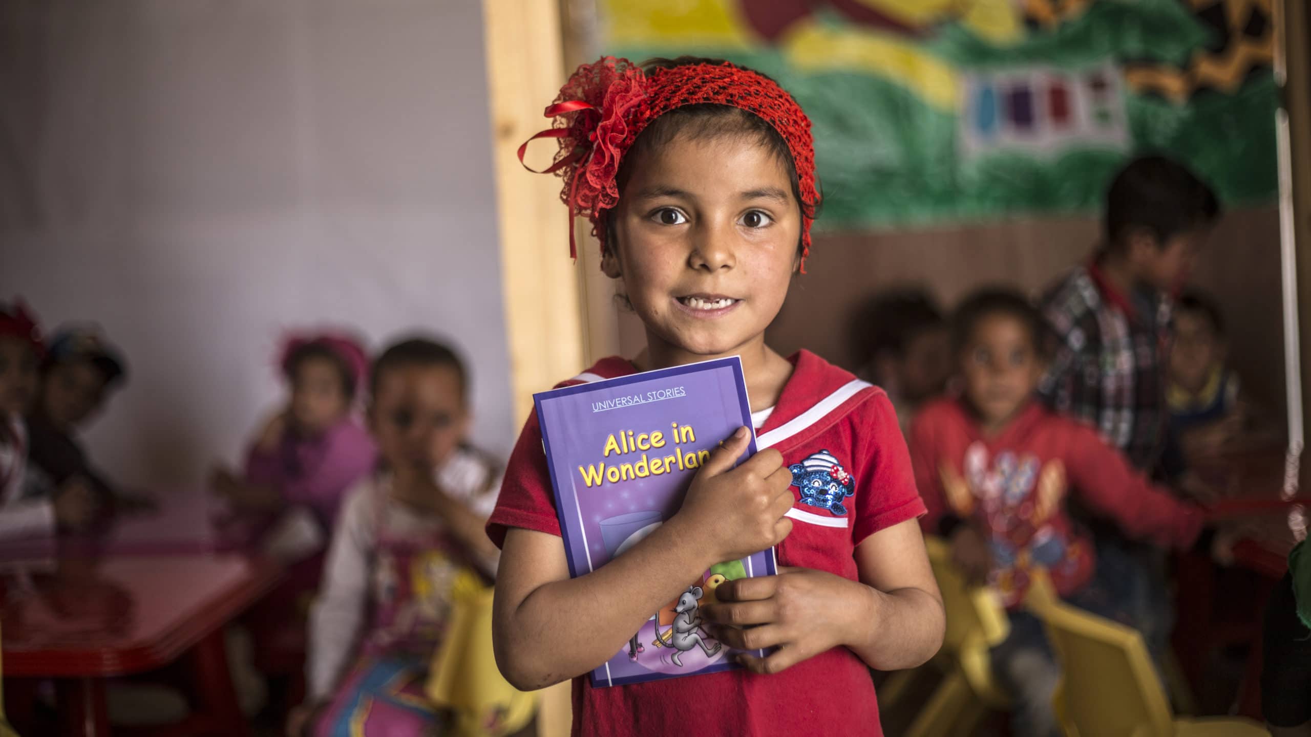 EI jente med rødt hårbånd står og holder en lærebok foran et klasserom