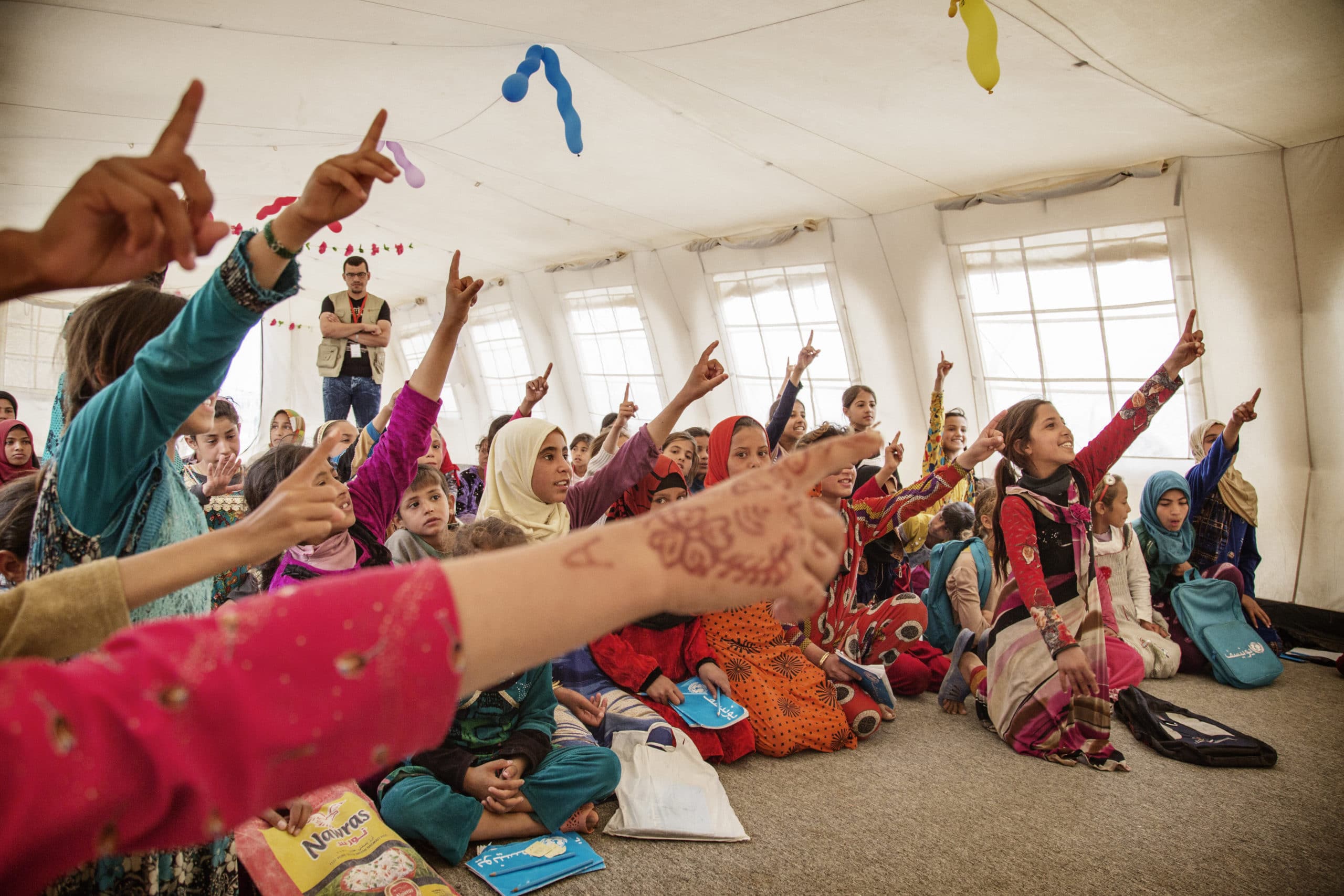 En gruppe barn sitter i et telt i en flyktningleir og rekker opp hånden i undervisningen