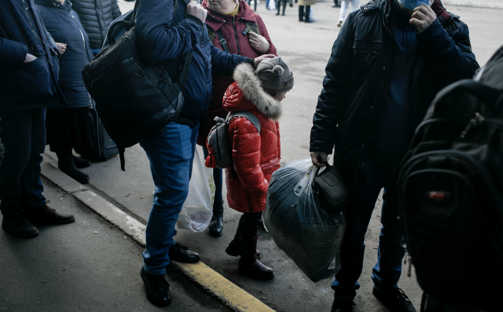 En jente i rød jakke står sammen med en flokk voksne og venter på en buss som skal ta dem ut av Kyiv.