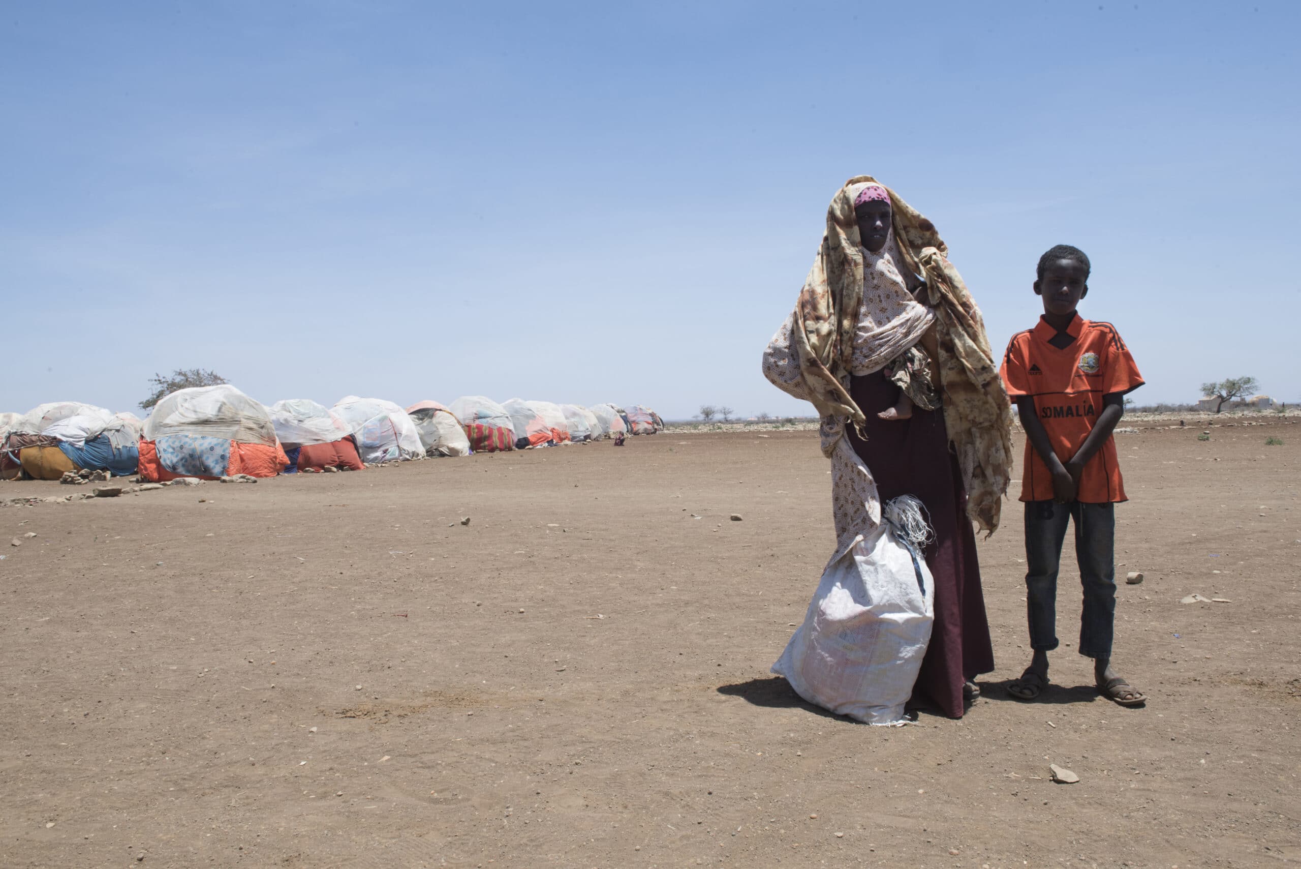 En mor står sammen med 2 av sine sønner og eiendeler i en pose, foran en flyktningleir i Somalia.