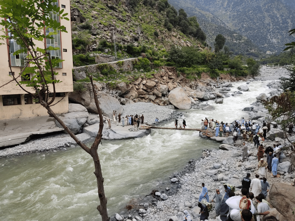 En flokk med mennesker bærer med seg sine eiendeler og krysser en oversvømt elv i Pakistan.