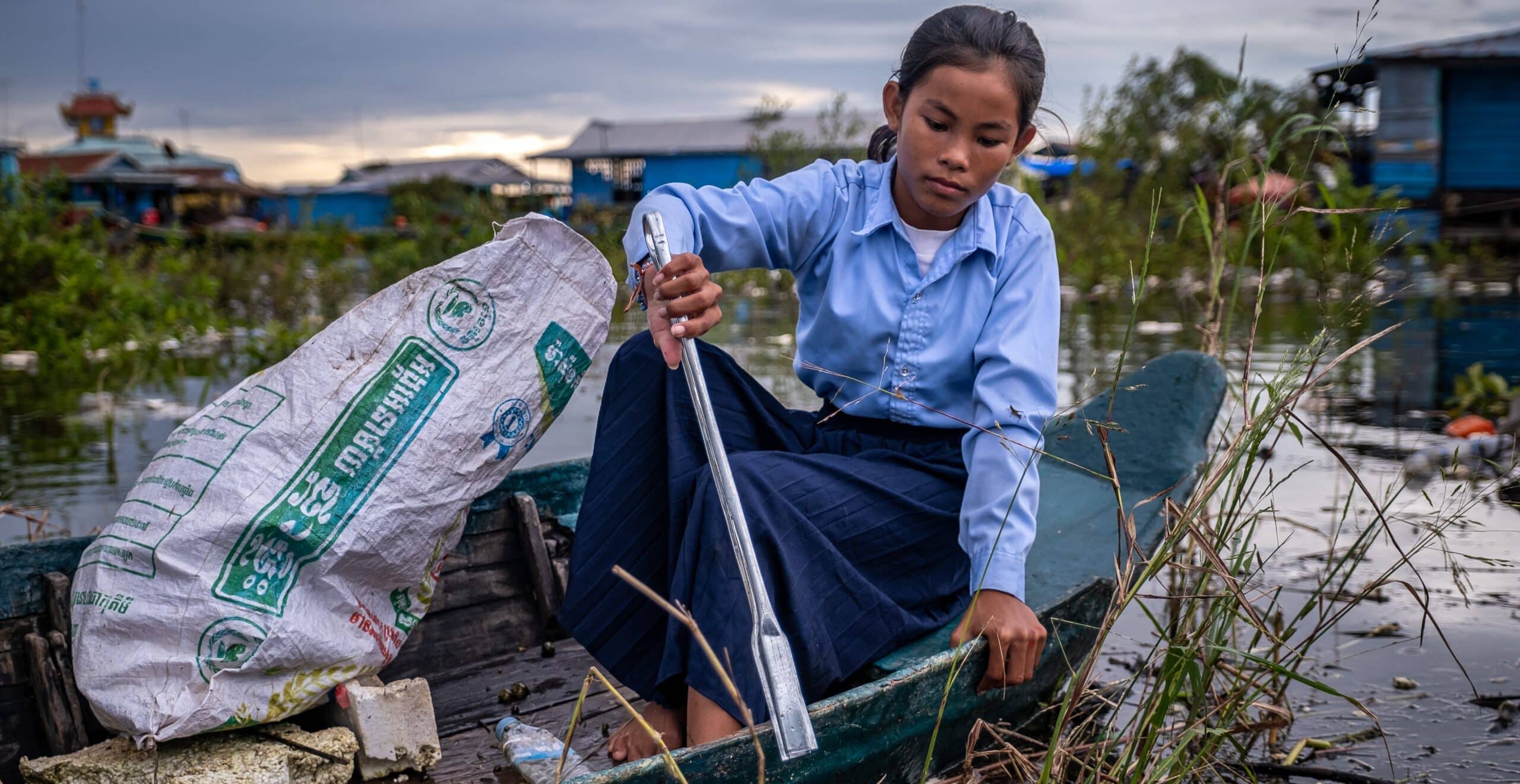 En jente sitter i en trebåt og plukker søppel opp av vannet med en tang