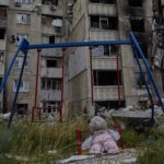 Et ødelagt huskestativ med en forlatt bamse står tomt foran et ødelagt boligkompleks