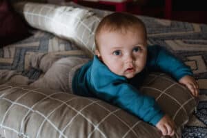 En baby i blå genser og grå bukse ligger på magen på en pute og kikker i kamera