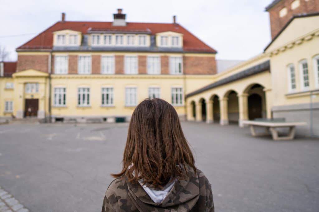 Ei jente står med ryggen til kamera og ser utover skolegården og mot en gul skole.