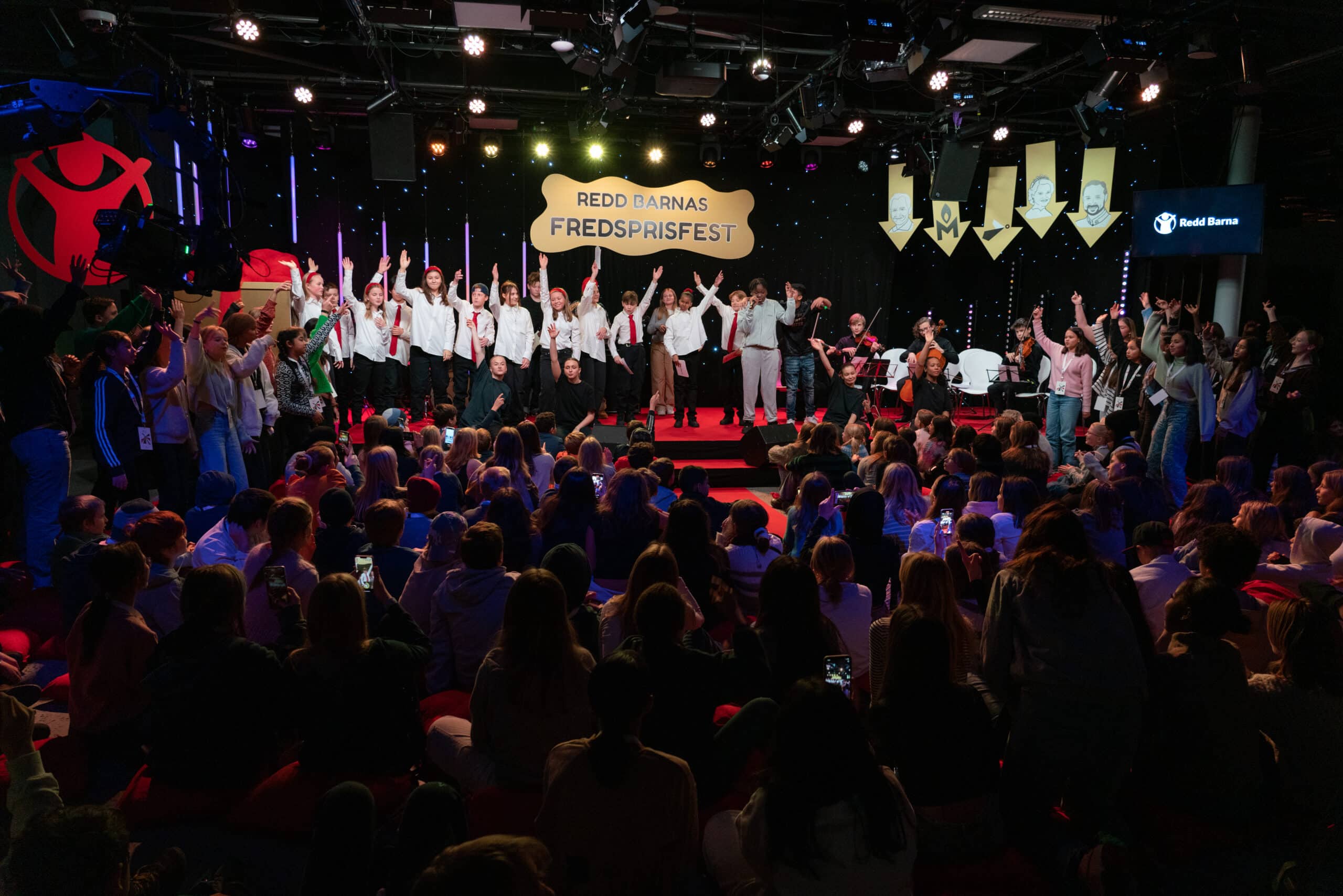Bilde fra Redd Barnas fredsprisfest 2023. Mange barn i salen og tolv elever i festkomiteen på scenen sammen med artister.