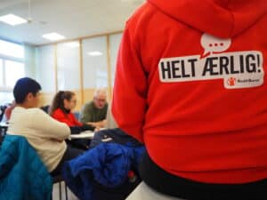 Et klasserom med elever som jobber og en person fra prosjektet om medvirkning, Helt Ærlig, i forgrunnen med rød hettegenser på.
