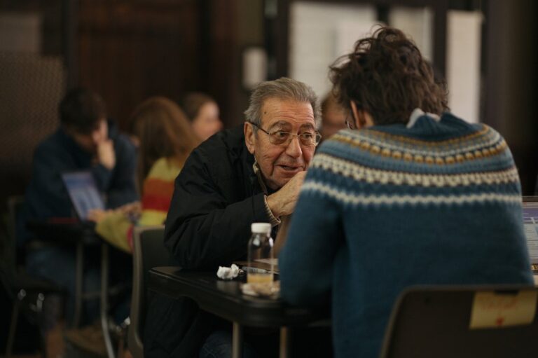 En eldre mann med briller sitter dypt engasjert i en samtale med en yngre person.