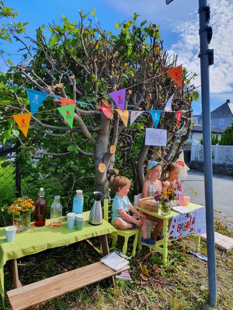 Tre barn som sitter foran et bord. De holder basar og det er sommer.