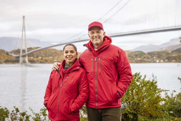 Tom Stiansen og Jorun Stiansen står i røde jakker foran vann og en bro