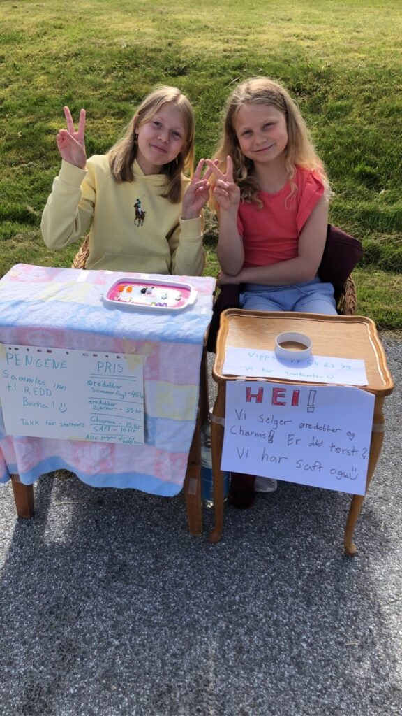To jenter sitter foran et bord, de holder basar. De selger øredobber de har laget. De holder fingrene oppe og viser peace tegn.