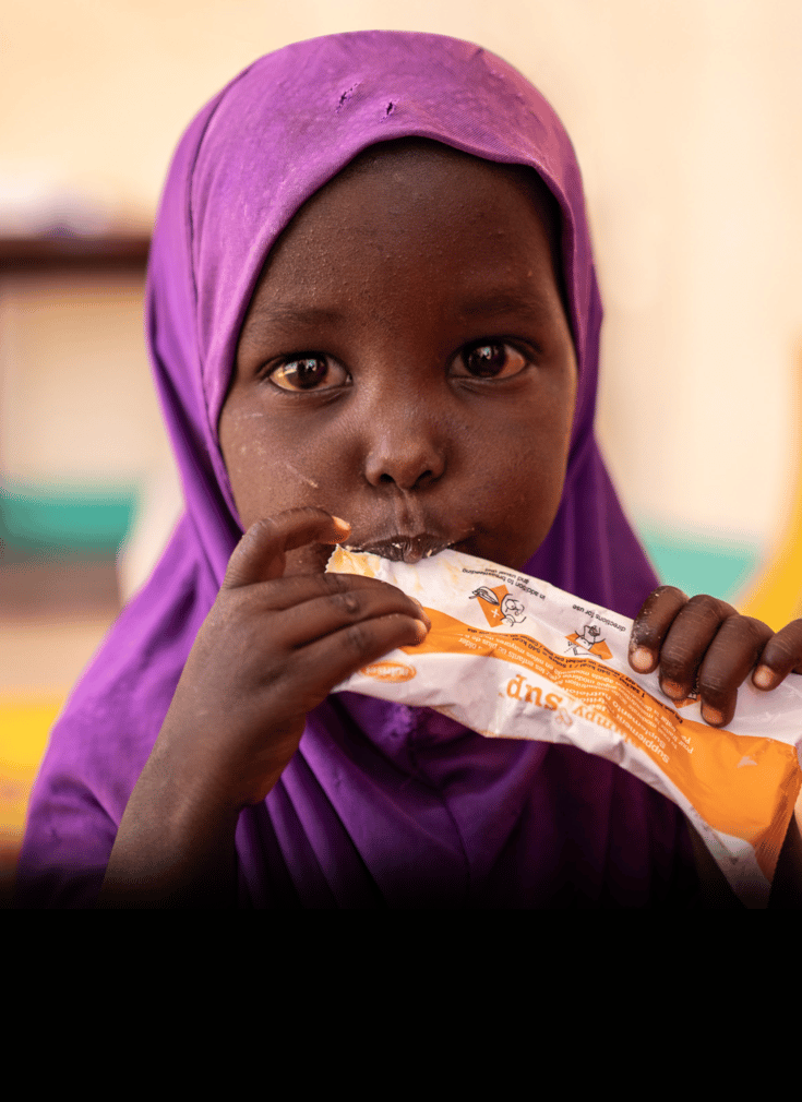 Jente fra Somalia med lilla hijab som ser inn i kameraet og spiser på Redd Barnas peanøttgrøt.