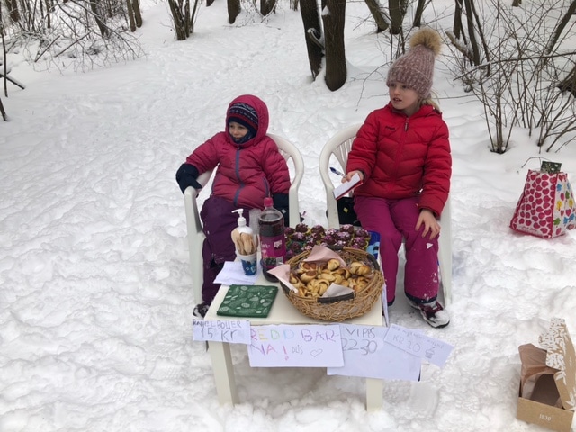 To jenter sitter foran et bord, de har på vintertøy og det er masse snø rundt dem. De er inne i en skog. Det er saft og sjokolademuffins og kanelboller på bordet.