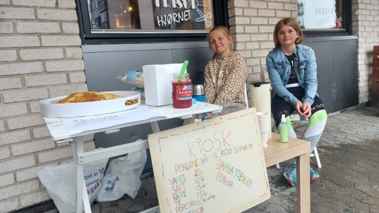 To barn som selger vafler, de smiler inn i kamera. De har laget en plakat der det står kiosk og hva de ulike tingene de selger koster.
