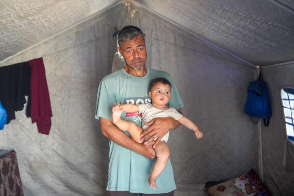 Raheem står og holder datteren sin i teltet i Gaza, mens han ser ned i bakken.