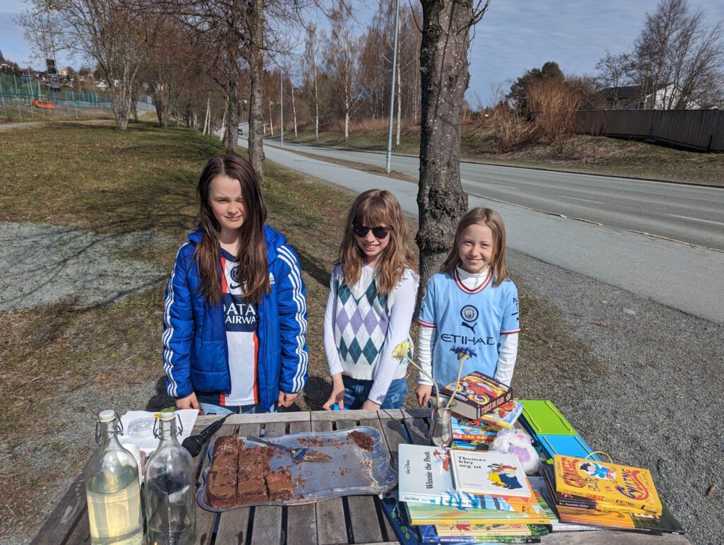 Tre fornøyde jenter holder en basar ute i solen og selger kake, saft og gamle leker de ikke lenger bruker.