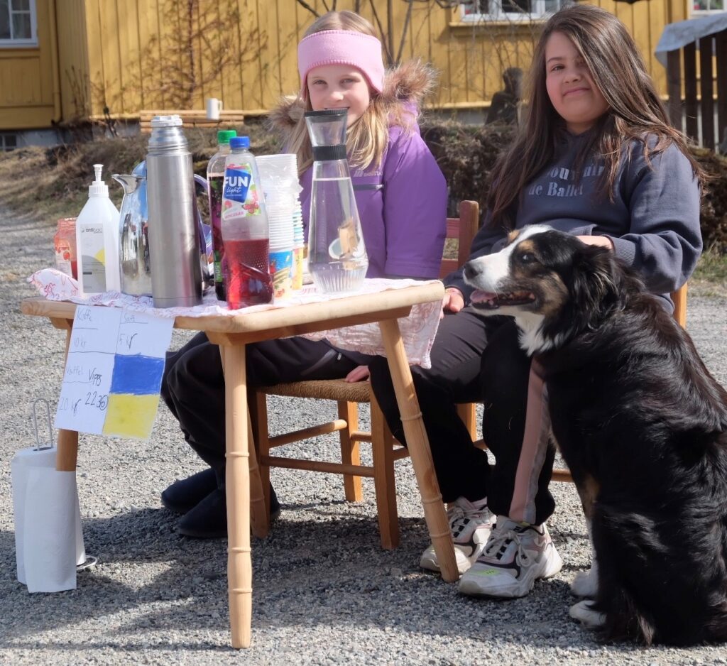 To jenter og en hund sitter foran et bord. De selger vafler, saft og kaffe.