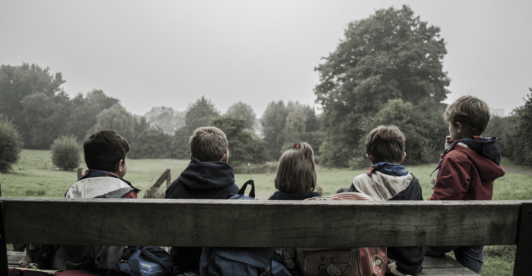 5 barn sitter på en trebenk i skogen med ryggen til