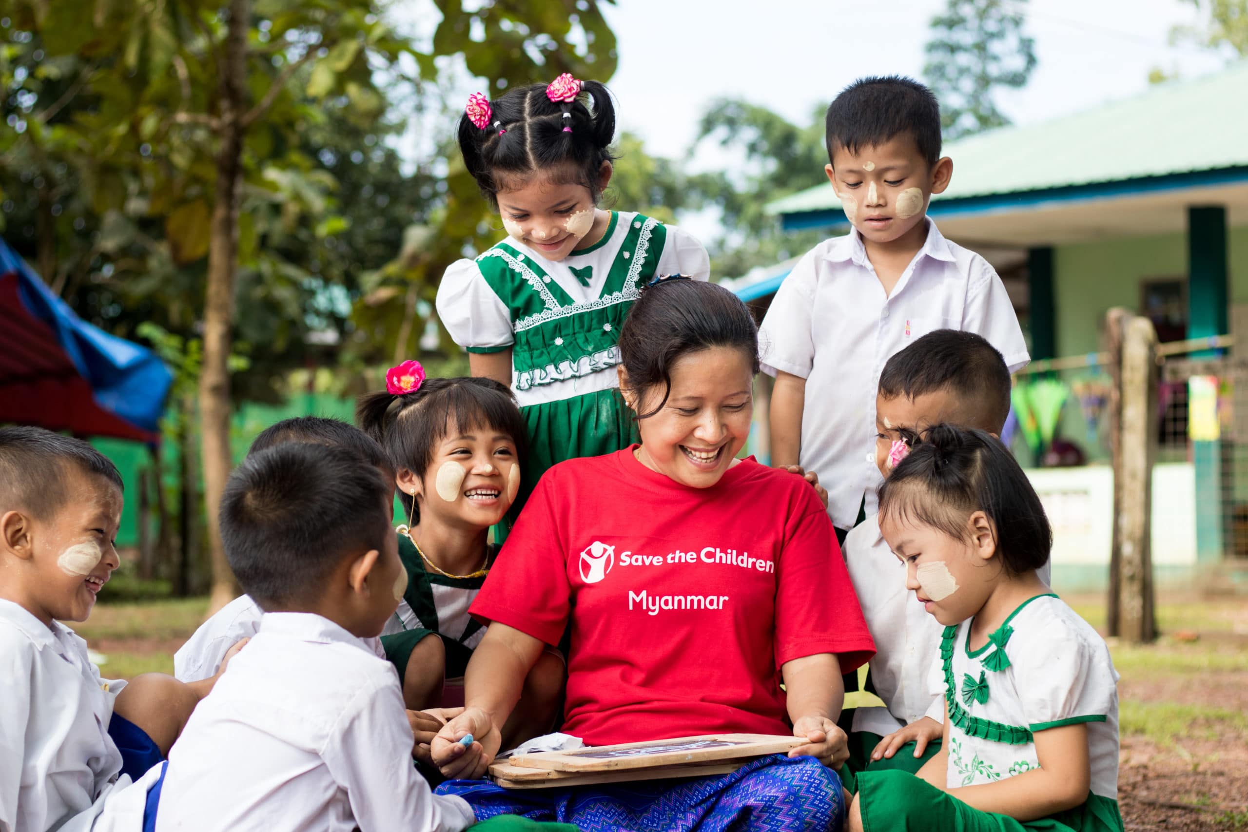 Flere glade barn står rundt en lærer med Redd Barna t-skjorte.