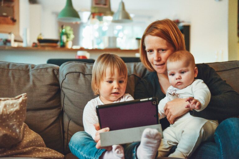 En mamma med to et lite barn og en baby som sitter i en sofa og ser på en ipad