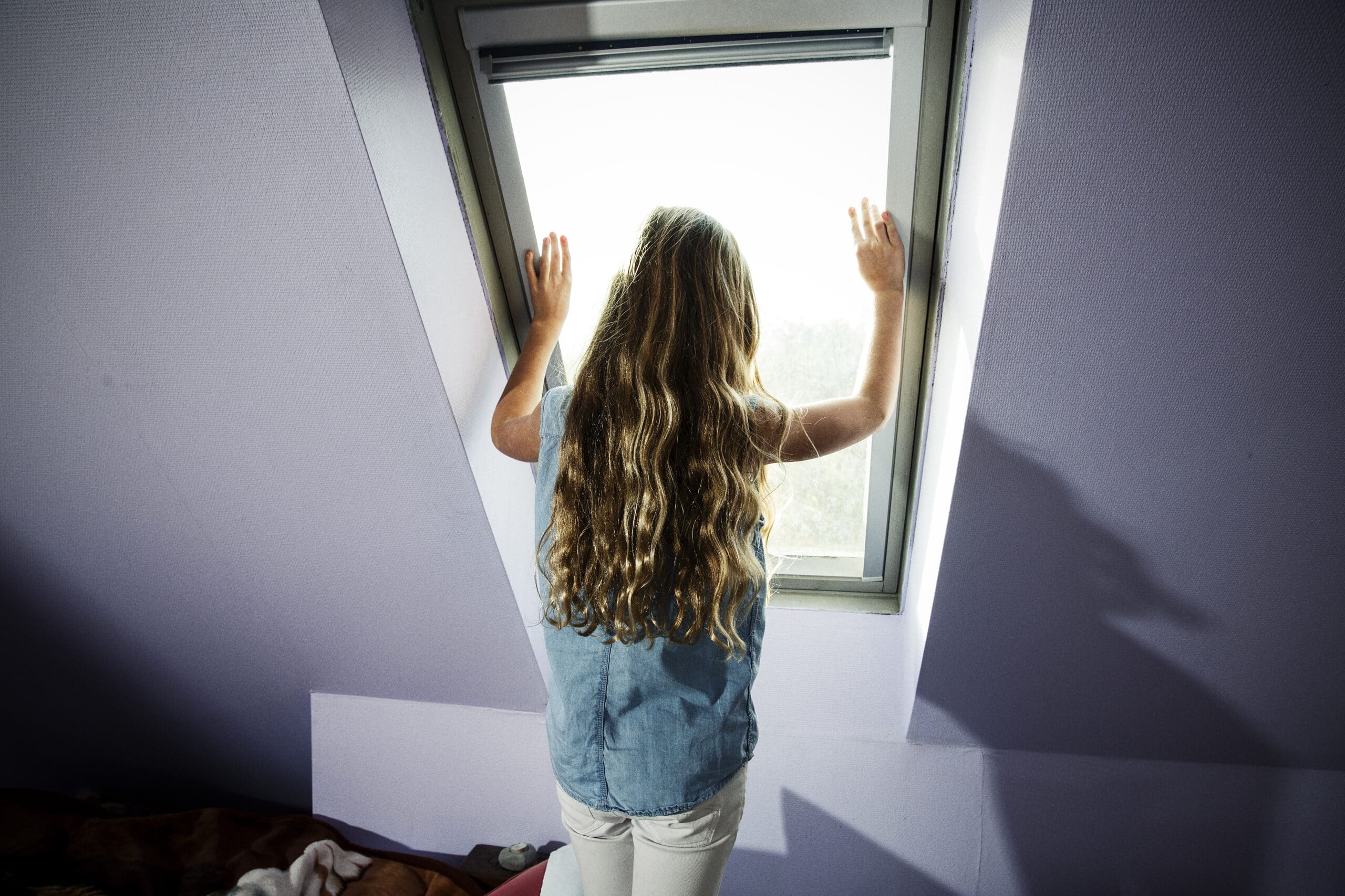En jente med langt krøllete hår står og kikker ut av et loftsvindu