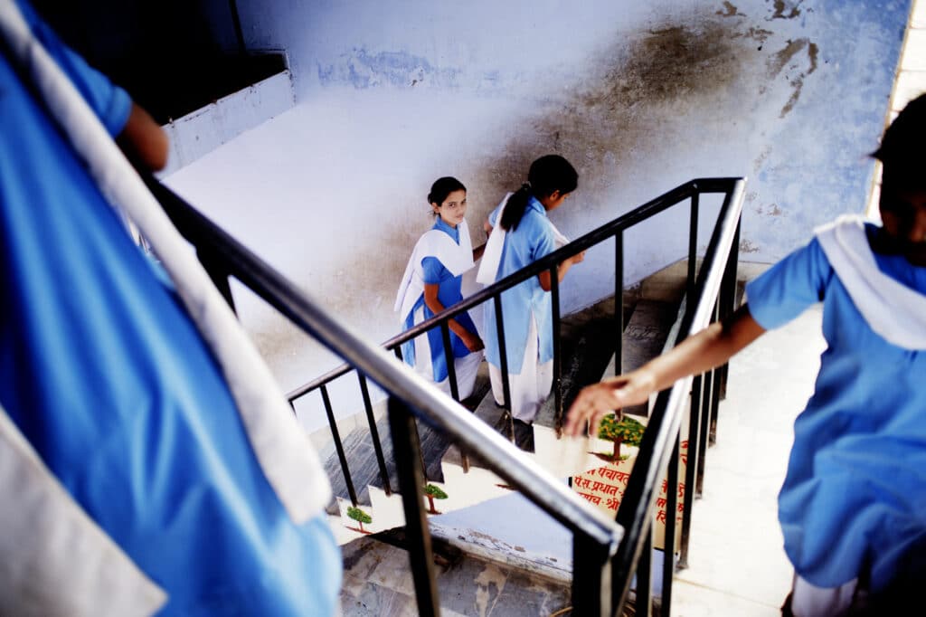 To jenter som går opp en trapp i et skolebygg i India. Den ene jenten ser i kamera. På trappen er det malt trær. Veggene er gamle og slitte. 