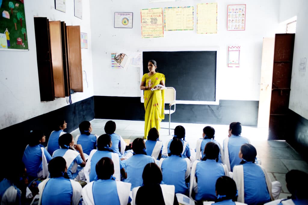 En indisk kvinne i gul drakt underviser et klasserom med jenter ikledd blått og hvitt. De sitter i et klasserom.