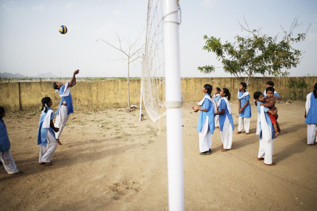 Jenter i blå og hvite klær spiller volleyball. Ballen er høyt i luften og en jente hopper og strekker seg etter ballen.