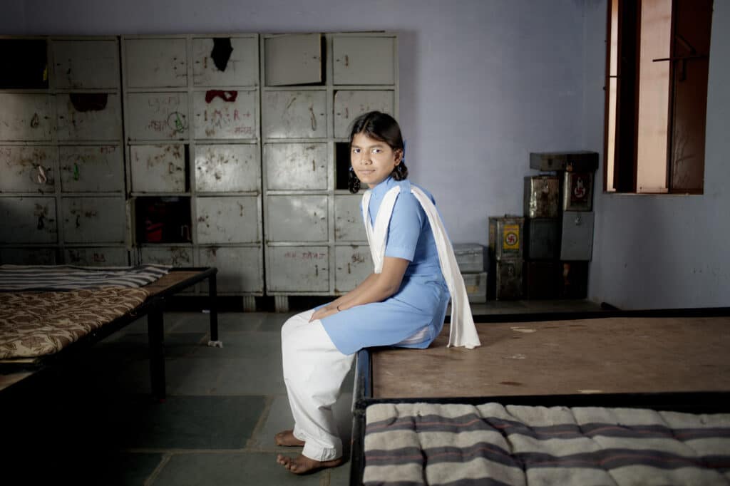 En indisk jente sitter på en seng og ser i kamera. Hun har på seg blå og hvite klær.
