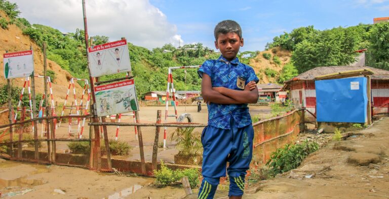 En gutt fra Bangladesh ikledd blå klær ser i kamera med armene i kryss. I bakgrunnen er en inngjerdet lekeplass