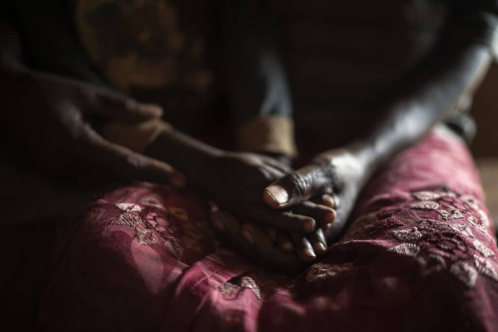En afrikansk kvinne holder hendene til barnet sitt. Hendene hviler i fanget til en rød-rosa kjole.