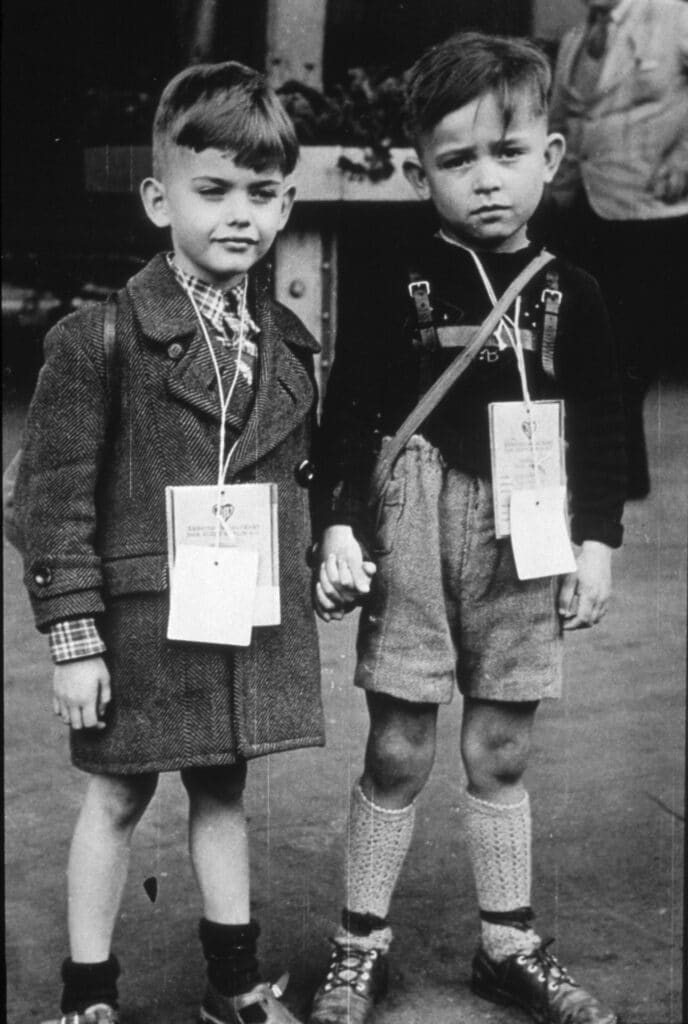 Historisk sort-hvitt-foto av to små gutter med navnelapper og sekker som holder hender.