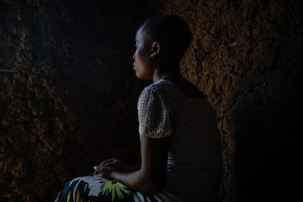En ung afrikansk jente sitter med ryggen til. Hendene er knyttet i fanget. Hun sitter i en gjørmehytte.