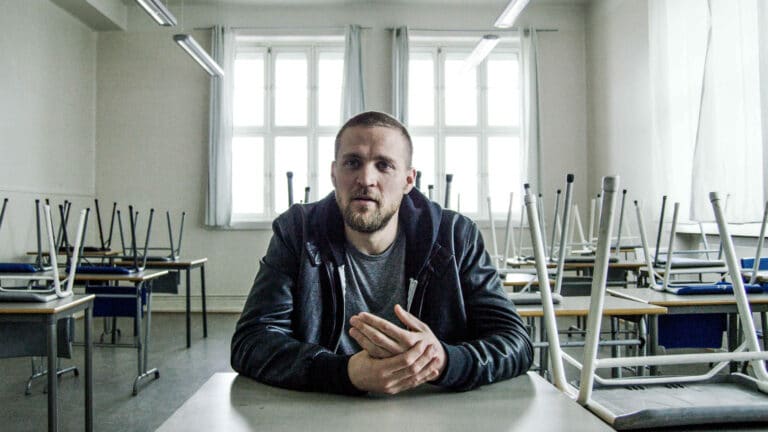 Foto av skuespiller Tobias Santelmann som sitter i et klasserom med hettegenser og skinnjakke. Illustrasjonsfoto fra Redd Barna film.