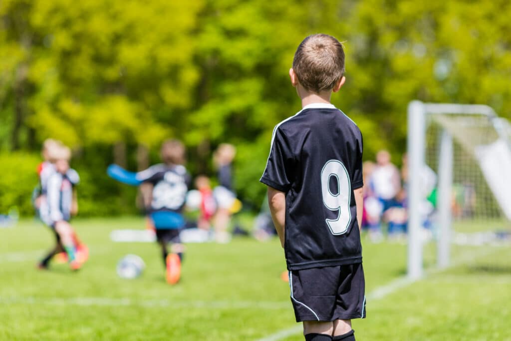 En gutt står ved en fotbbalbane med fotballdrakt. Han står på sidelinjen og ser på de andre spille fotball.