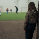 En jente står med ryggen til og lener seg opp mot et tre mens hun ser på andre barn spille fotball