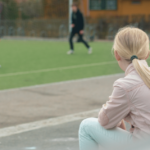 En jente sitter i rosa jakke og hvite bukser ved en fotballbane med ryggen til. Hun ser bort på andre barn og unge som spiller fotball.