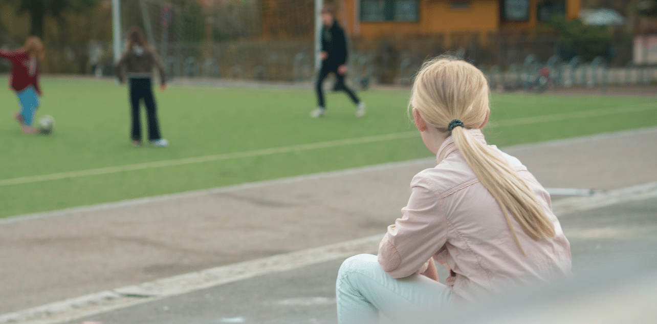 En jente sitter i rosa jakke og hvite bukser ved en fotballbane med ryggen til. Hun ser bort på andre barn og unge som spiller fotball.