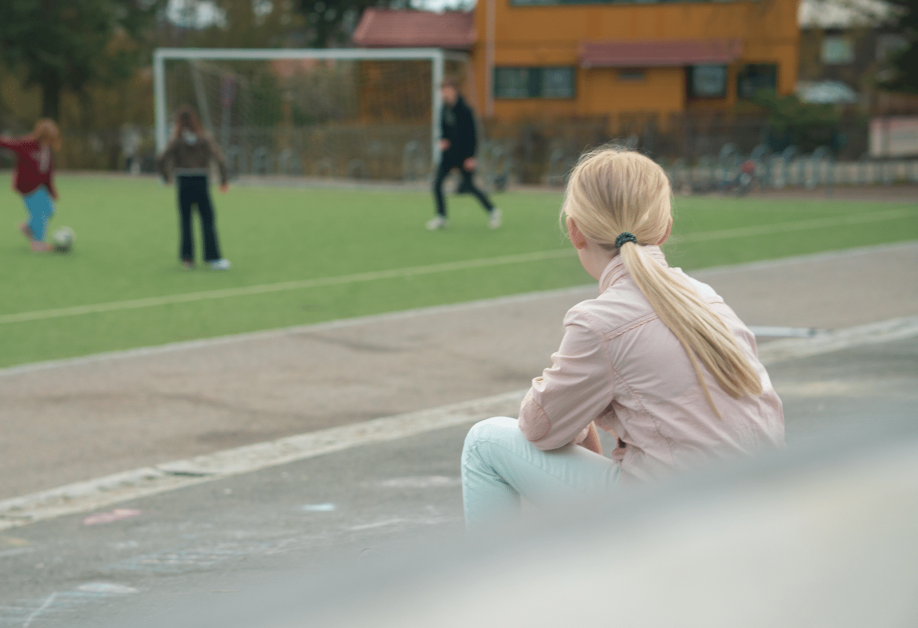 En jente sitter i rosa jakke og hvite bukser ved en fotballbane med ryggen til. Hun ser bort på andre barn og unge som spiller fotball.